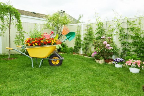A well-maintained backyard garden in Wandsworth featuring a vibrant green lawn with evenly cut grass, bordered by flower beds filled with blooming pink and purple flowers. The garden is enclosed by a white wooden fence covered with climbing green vines and leafy foliage. In the foreground, a yellow wheelbarrow is filled with colourful flower pots, gardening tools, and decorations including a small pink flamingo ornament. The background shows a tall, leafy shrub or small tree providing some height variation. The overall scene depicts a tidy, landscaped outdoor space suitable for gardening services offered in Wandsworth, with natural daylight and a slightly overcast sky adding soft lighting to the scene.