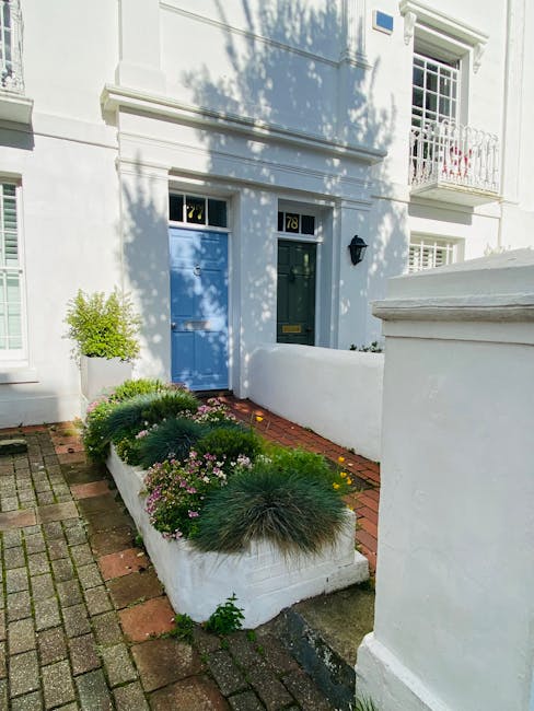 A charming front garden featuring a narrow pathway paved with pebble stones leading to a black front door set into a cream-colored house exterior. The garden is bordered by a low brick wall and black 